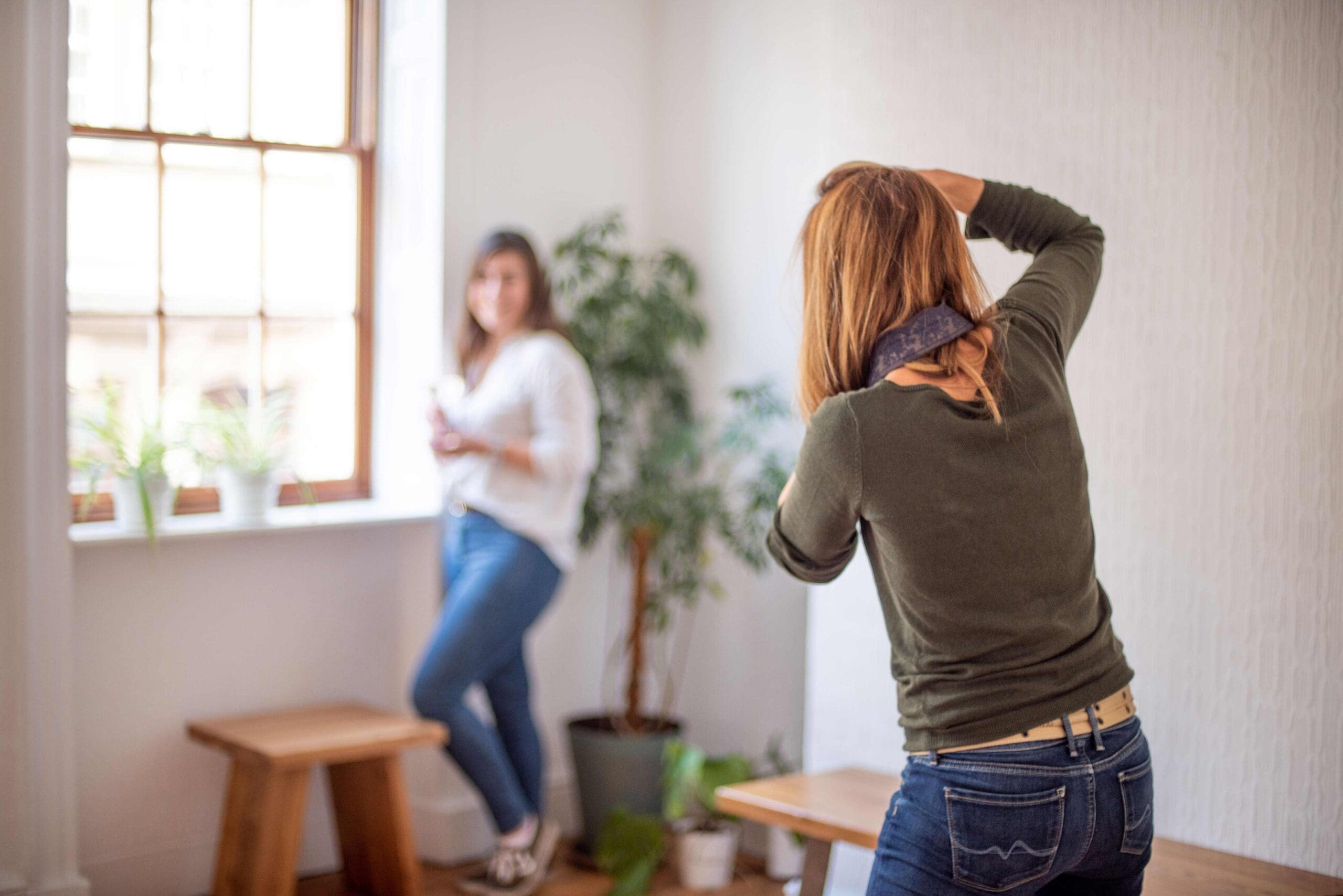 An image from behind of a lady with long blonde brown hair photographing another woman. The photographer wears a dark green long sleeve top over blue jeans The other lady is out of focus in the distance stood by a window smiling to camera. She wears a white shirt over blue jeans.