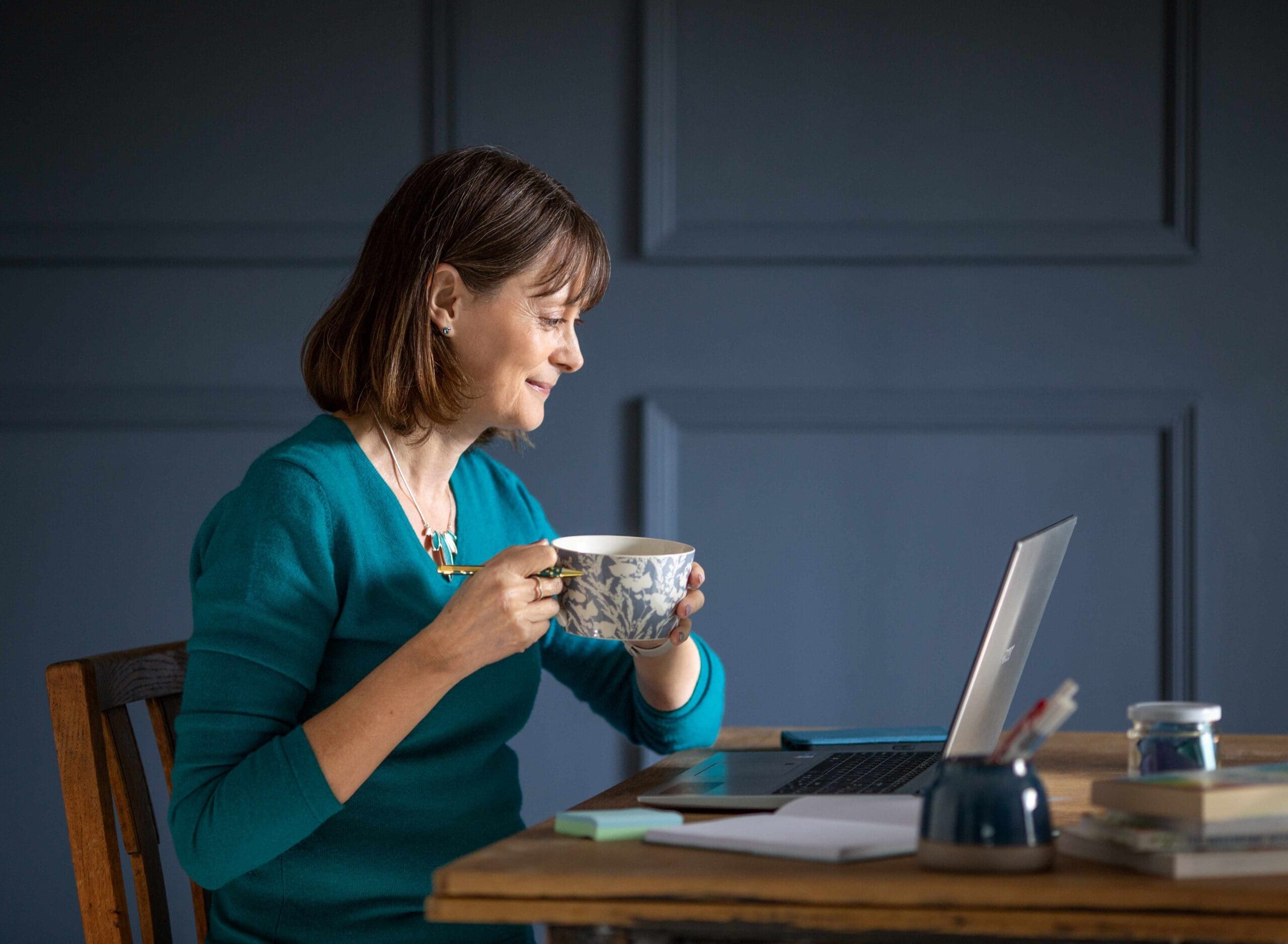 A woman with short dark hair wearing a teal jumper sits at a desk holding a cup of tea. She is looking at the laptop open on the desk in front of her with a slight grin. In the background is a grey pannelled wall.