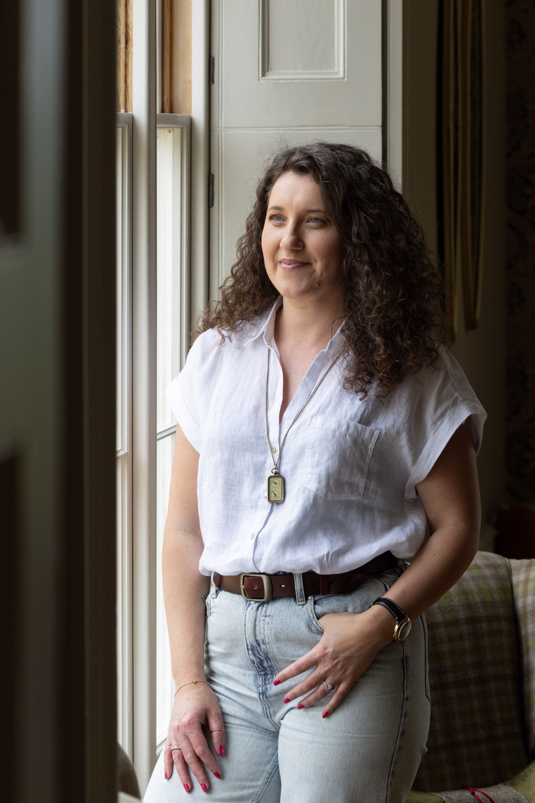 An image of a woman with long curly dark hair leans in by a window. She wears a short sleeve white shirt over light blue jeans with a dark belt. She looks out of the window and has one thumb yucked in a pocket casually.